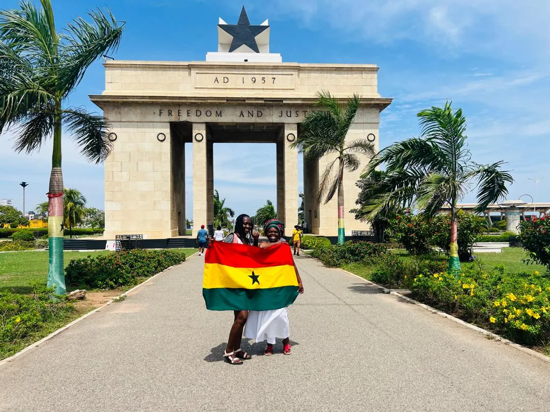 Two travellers holding a Ghana flag in front of Independence Arch in Accra