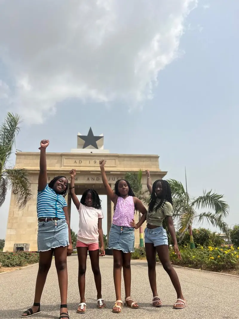 Four girls raising their arms in front of Independence Arch in Accra