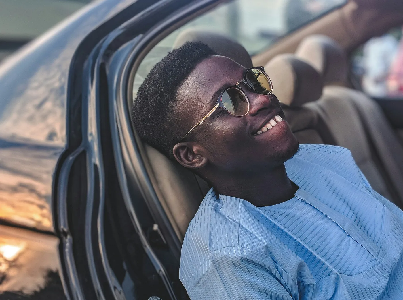 A smiling young man in sunglasses and a light blue shirt relaxes in the open doorway of a car